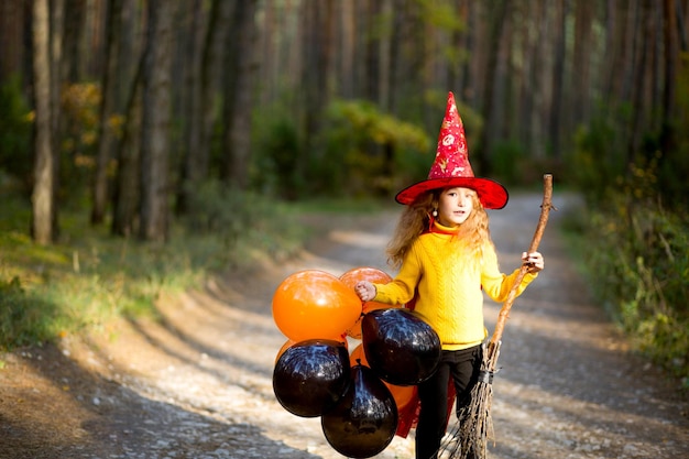 Una ragazza con un costume da strega e un cappello su una scopa con palloncini arancioni e neri sta giocando nella foresta autunnale andando a una festa di Halloween