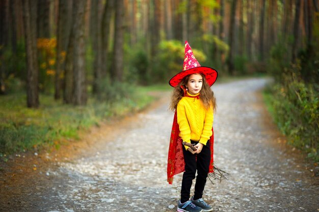 Una ragazza con un costume da strega e un cappello su una scopa sta giocando nella foresta autunnale andando a una festa di Halloween