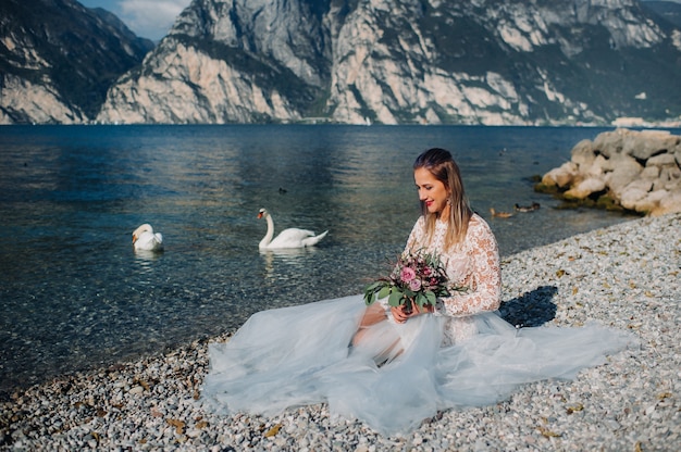 Una ragazza con un elegante abito bianco è seduta sull'argine del Lago di Garda, mentre una donna è fotografata sullo sfondo di una montagna e di un lago in Italia.Torbole.