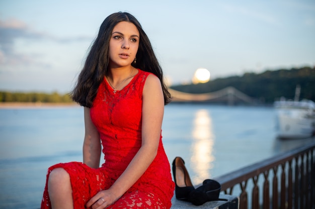 Una ragazza con un lungo vestito rosso è seduta sul recinto dell'argine del fiume.