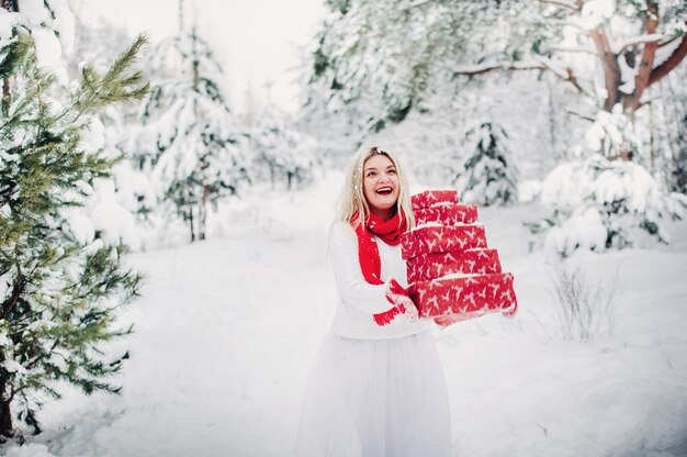 Una ragazza con un maglione bianco con regali di Natale in mano si trova in una foresta innevata. Donna con regali di Natale fuori in inverno