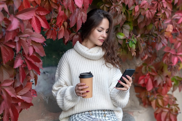 Una ragazza con un maglione bianco guarda il telefono e tiene un bicchiere.