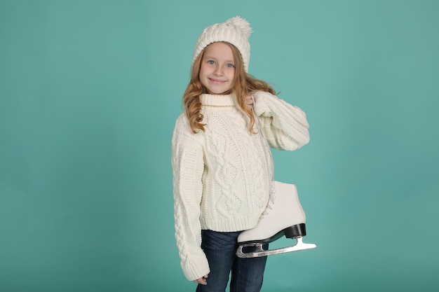 una ragazza con un maglione bianco lavorato a maglia e un cappello bianco tiene in mano dei pattini da pattinaggio artistico bianchi