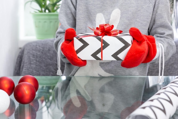 Una ragazza con un maglione grigio con un coniglio tiene in mano un regalo di Natale con guanti rossi nelle sue mani nuovo y...