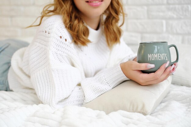 Una ragazza con un maglione lavorato a maglia bianco giace su un letto con una tazza di bevanda calda in mano sullo sfondo di un muro di mattoni bianchi