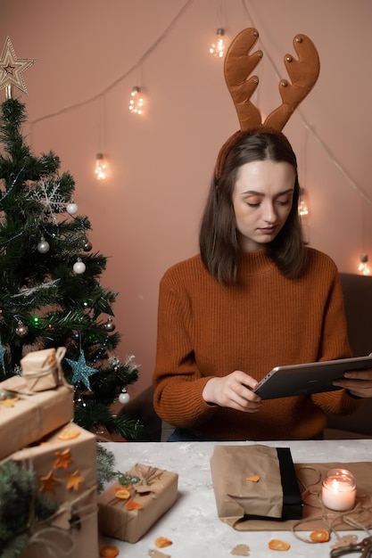 una ragazza con un maglione marrone tiene in mano un tablet e impacchetta i regali per il nuovo anno