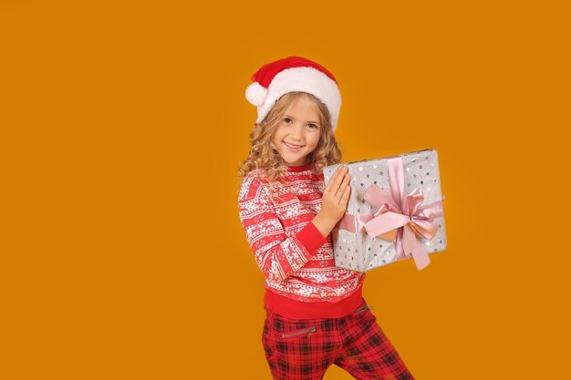 una ragazza con un maglione natalizio in pantaloni a quadri e un cappello di Babbo Natale tiene una scatola regalo