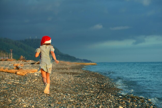 Una ragazza con un vestito a righe e un cappello rosso di Babbo Natale ci corre dando le spalle lungo la riva del mare