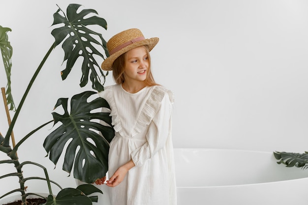 una ragazza con un vestito bianco e un cappello di paglia in una stanza con piante e un bagno