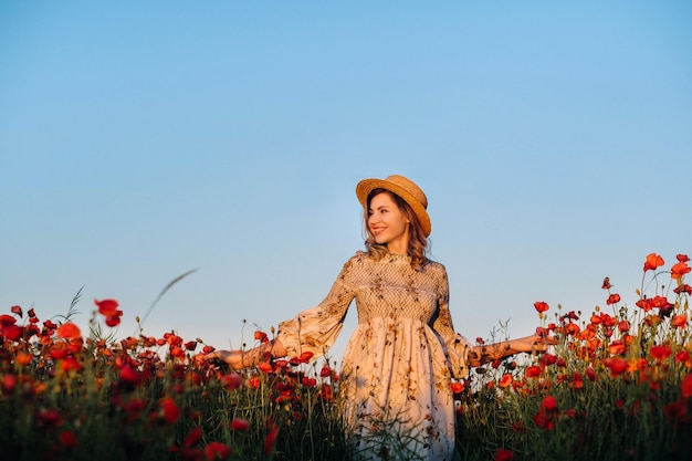 Una ragazza con un vestito con un cappello cammina in un campo con papaveri al tramonto