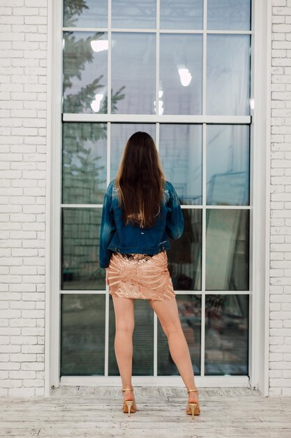 Una ragazza con un vestito corto e una giacca di jeans sta con la schiena vicino alla finestra