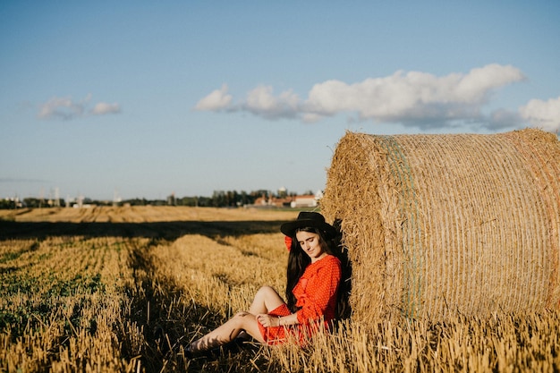 Una ragazza con un vestito rosso e un cappello nero è seduta vicino a un pagliaio sotto il sole della sera