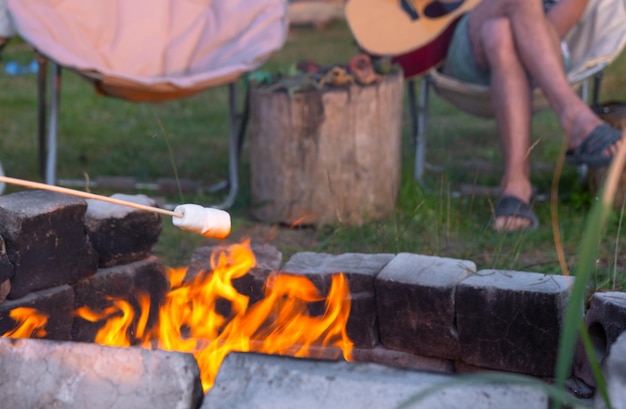 Una ragazza con una camicia a quadri arrostisce marshmallows su un fuoco nel cortile della casa il padre suona la chitarra la sera la famiglia si riunisce al fuoco del campo picnic all'aperto in sedie turistiche