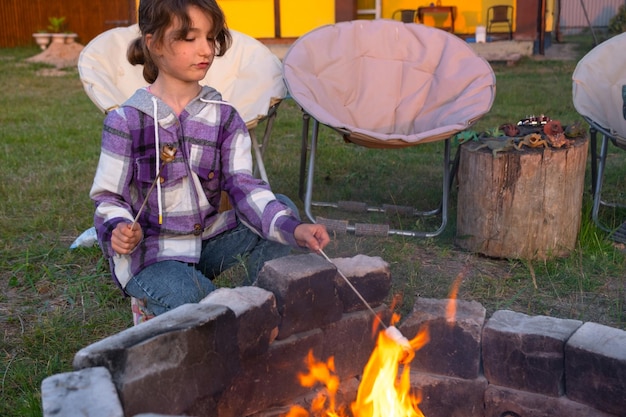 Una ragazza con una camicia a quadri arrostisce marshmallows su un fuoco nel cortile della casa La famiglia della sera si riunisce accanto al fuoco del campo sedie turistiche all'aperto