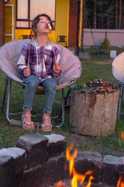 Una ragazza con una camicia a quadri arrostisce marshmallows su un fuoco nel cortile della casa La famiglia della sera si riunisce accanto al fuoco del campo sedie turistiche all'aperto