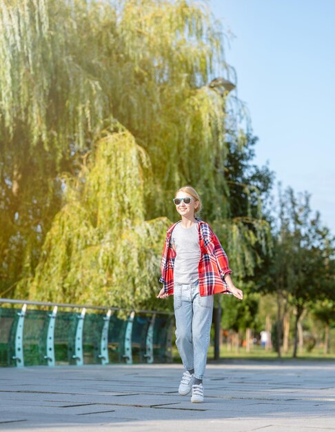 Una ragazza con una camicia a quadri salta e corre lungo un sentiero in un parco cittadino in una soleggiata giornata estiva.