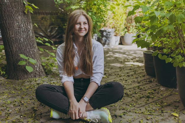 Una ragazza con una camicia bianca si siede vicino ad alberi e foglie