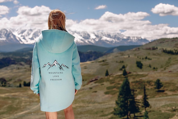 Una ragazza con una felpa oversize blu con una stampa è in piedi con la schiena e guarda un bellissimo paesaggio di foresta di montagna e un cielo soleggiato con diverse nuvole.