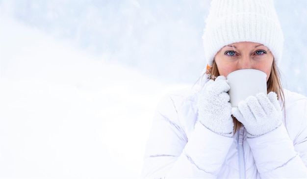 Una ragazza con una giacca bianca e un cappello bianco tiene in mano una tazza di tè durante una nevicata sullo sfondo di bellissimi cumuli di neve