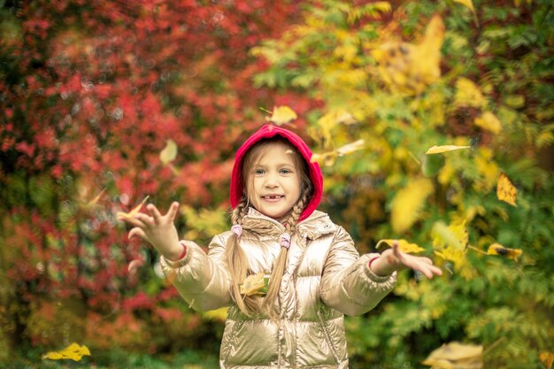 Una ragazza con una giacca color oro sorride lanciando foglie rosa e gialle in una giornata autunnale