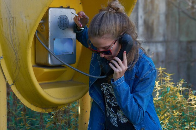 Una ragazza con una giacca di jeans blu occhiali da sole e con una sigaretta in una cabina telefonica gialla