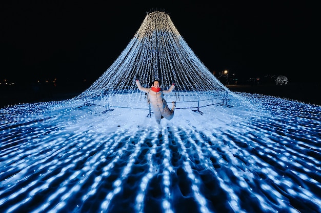 Una ragazza con una giacca grigia in inverno con le luci della sera accese sulla strada di Natale