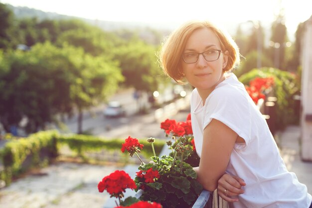 Una ragazza con una maglietta bianca e occhiali sta sul balcone e gode della bellezza e della cura. luce del sole brillante.