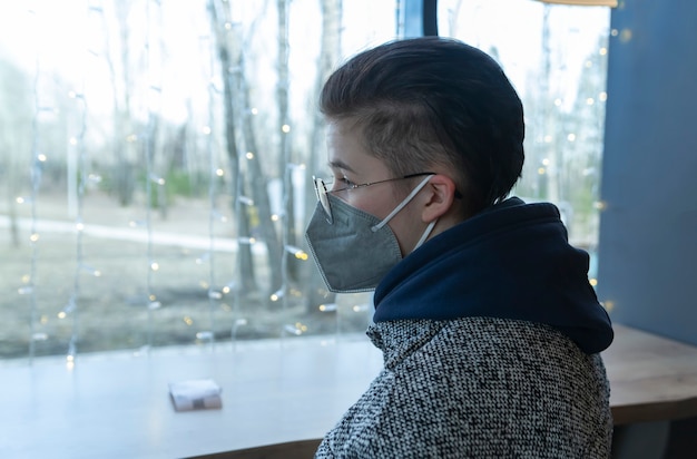 Una ragazza con una maschera medica si siede in un caffè e guarda tristemente fuori dalla finestra