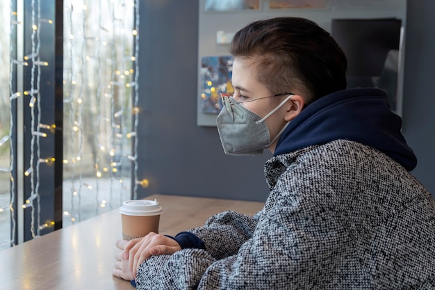 Una ragazza con una maschera medica si siede in un caffè e guarda tristemente fuori dalla finestra