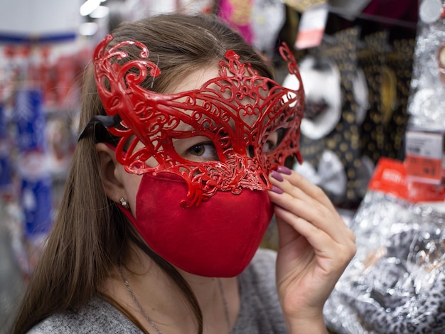 Una ragazza con una maschera protettiva rossa prova una maschera di carnevale in un negozio Lo sguardo dietro la maschera Acquistare decorazioni natalizie Ragazza con una maschera veneziana rossa La ragazza con due maschere