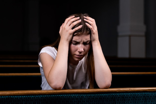 Una ragazza cristiana triste in camicia bianca è seduta e prega con umile cuore in chiesa