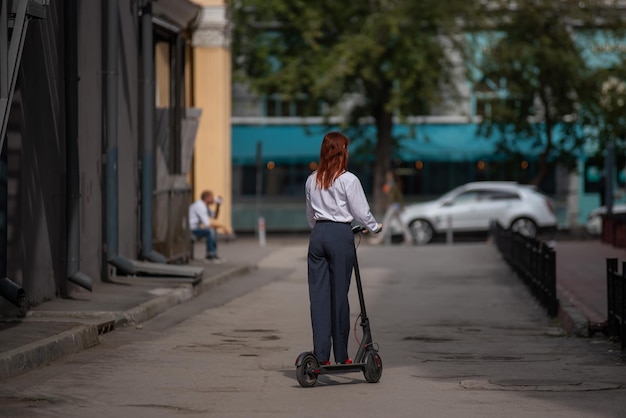 Una ragazza dai capelli rossi con una camicia bianca guida uno scooter elettrico lungo il muro. Una donna d'affari con un tailleur pantalone e tacchi alti rossi gira per la città con un moderno codice di abbigliamento da auto in ufficio