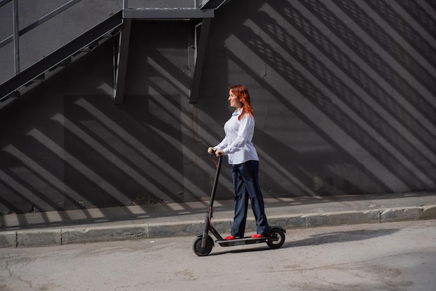 Una ragazza dai capelli rossi con una camicia bianca guida uno scooter elettrico lungo il muro Una donna d'affari in tailleur pantalone e tacchi rossi gira per la città in un'auto moderna Donna d'affari su uno scooter