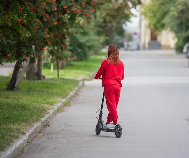 Una ragazza dai capelli rossi con una tuta da ginnastica rossa guida uno scooter elettrico Una giovane donna in abiti oversize gira per la città e ascolta musica usando le cuffie wireless Vista dal retro