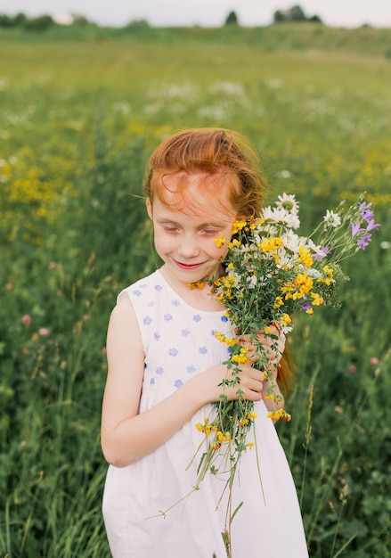 Una ragazza dai capelli rossi sta con un mazzo di fiori selvatici in mezzo a un campo.