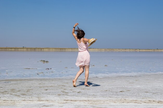 Una ragazza dai capelli scuri con un vestito rosa e un cappello di paglia corre a piedi nudi lungo le rive di un lago salato.