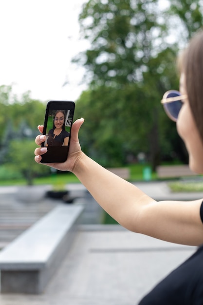 Una ragazza dalla pelle bianca in abiti moderni tiene in mano uno smartphone e parla su un collegamento video in un parco in estate.