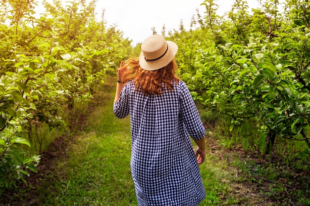 Una ragazza del giardiniere cammina attraverso un frutteto di mele verde in un vestito e un cappello blu