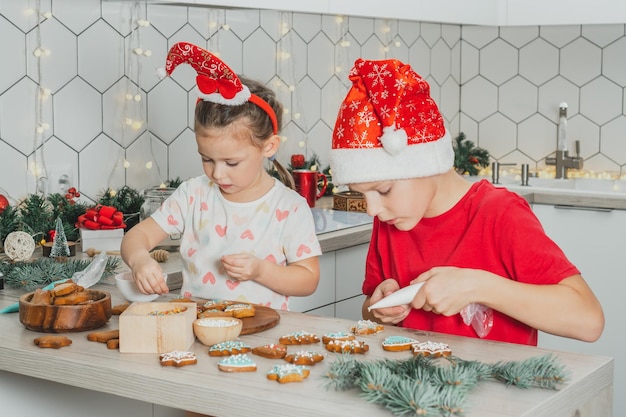 Una ragazza di 3 anni e un ragazzo di 8 anni con il cappello da Babbo Natale decorano i biscotti di pan di zenzero con glassa I fratelli nella cucina di Natale