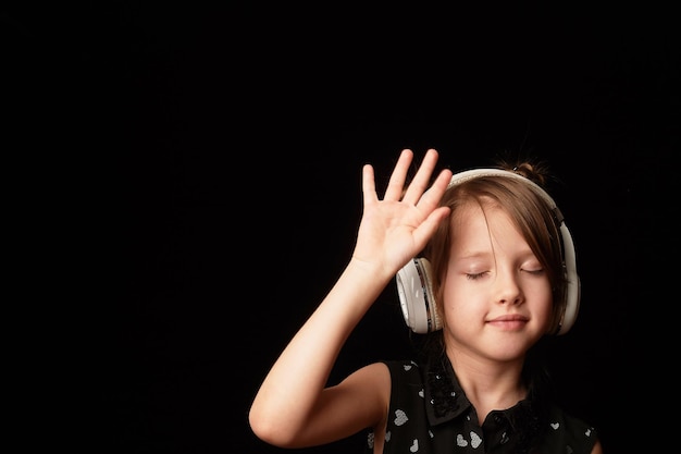 Una ragazza di 6 anni su uno sfondo scuro Ballando in cuffie wireless bianche Alza le mani Godendo la danza