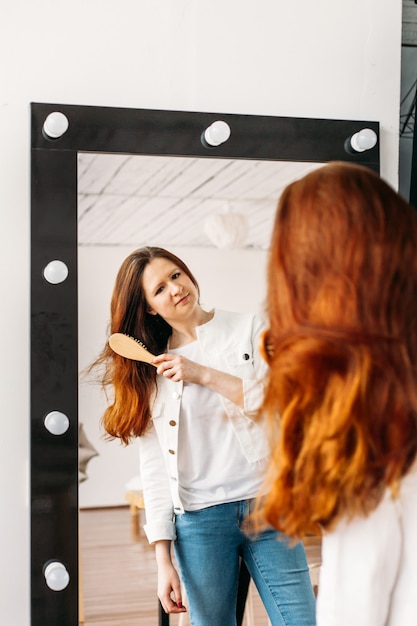 Una ragazza di aspetto europeo si trova di fronte a uno specchio, si pettina i capelli con un pettine, bellezza dei capelli, cura,