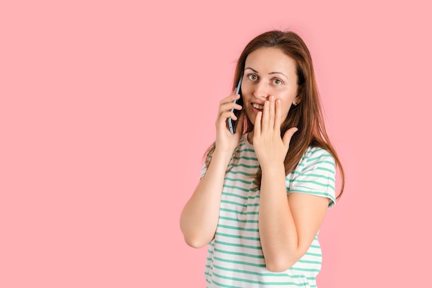 Una ragazza di mezza età in abiti casual sta parlando al telefono e si copre la bocca con il palmo per la sorpresa. Foto in studio su sfondo rosa