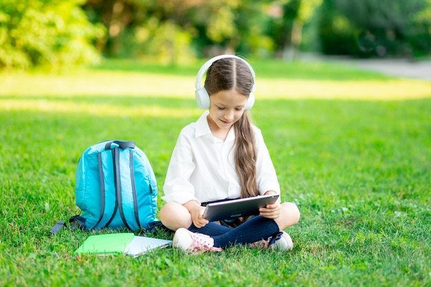 Una ragazza di prima elementare con uno zaino e un tablet con le cuffie su un prato verde legge un libro o fa i compiti torna a scuola