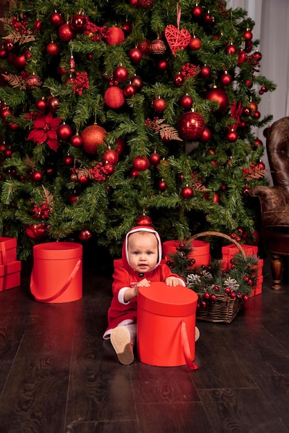 Una ragazza di un anno travestita Babbo Natale con regalo si siede sullo sfondo dell'albero di Natale decorato all'interno guardando la macchina fotografica