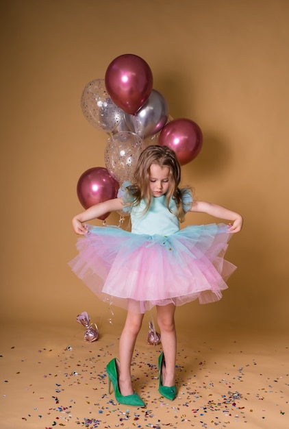Una ragazza divertente con un vestito festivo si trova nelle grandi scarpe di sua madre su uno sfondo beige con palloncini e coriandoli