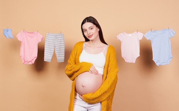 Una ragazza dolce, affascinante, gentile, con lunghi capelli scuri, in un cardigan lavorato a maglia giallo, con la pancia nuda, si tocca la pancia con le mani e sorride dolcemente, guarda dritto. Maternità, infanzia.