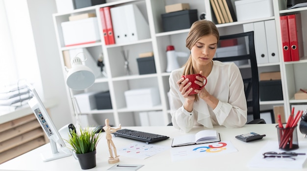 Una ragazza è seduta a un tavolo in ufficio, con in mano una tazza rossa e guardando i fogli con gli schemi.