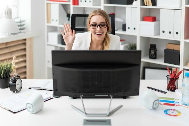 Una ragazza è seduta al tavolo in ufficio, guardando il monitor e agitando la mano.