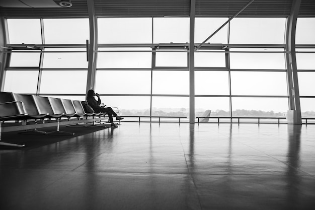 Una ragazza è seduta su una fila di sedili vuota davanti a una grande vetrata in un terminal di un aeroporto, in attesa di un volo.