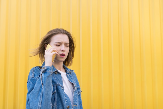 Una ragazza emotiva in giacca di jeans parla al telefono sullo sfondo del muro giallo.
